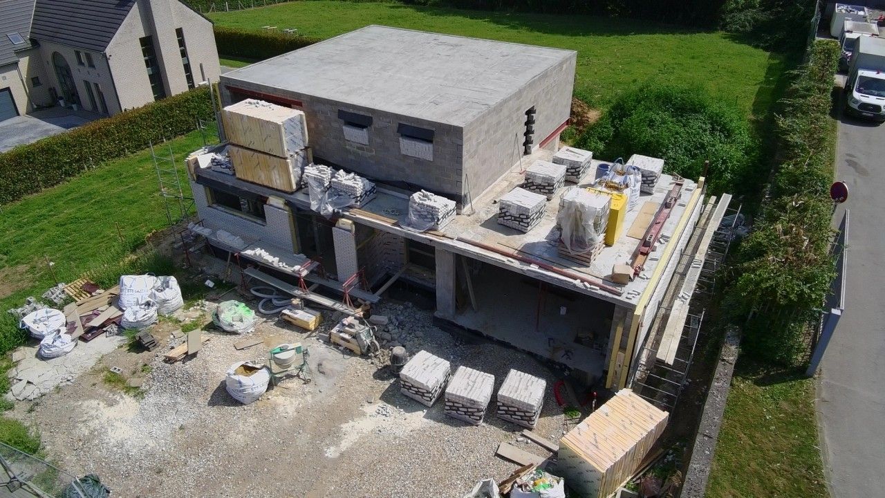 Vue aérienne du chantier d’une maison contemporaine neuve à Oupeye, montrant la toiture plate, les volumes en construction et la mise en œuvre du parement en briques claires.