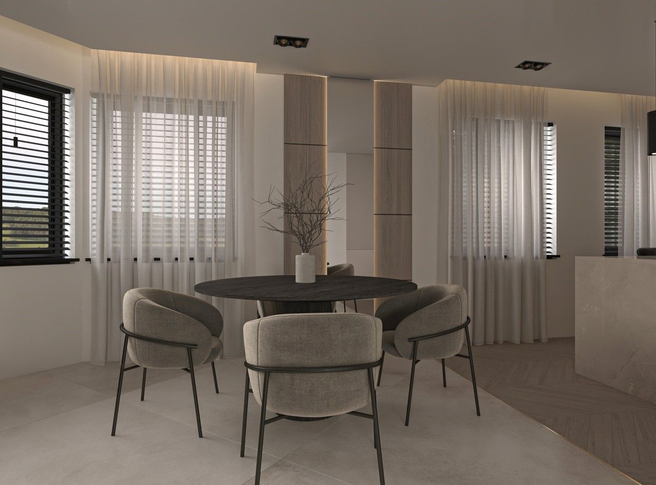 Vue de la salle à manger d’un appartement rénové à Liège, avec table ronde sombre, fauteuils rembourrés, grandes fenêtres habillées de voilages et menuiseries intégrées en bois clair.