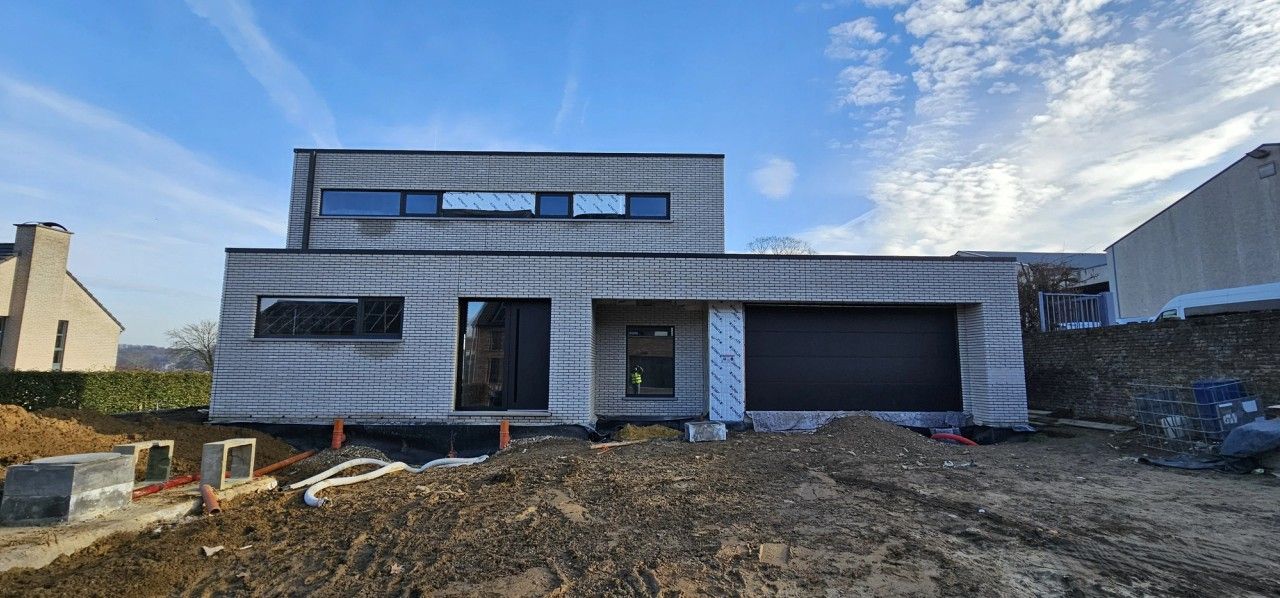 Chantier d’une maison contemporaine neuve à Oupeye montrant la façade avant en briques claires, le garage intégré et les châssis foncés en cours de finition.