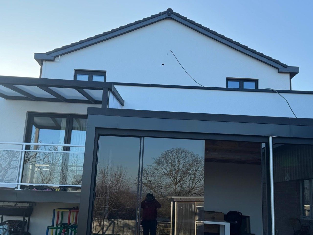 Vue de la façade arrière rénovée à Vottem, avec enduit clair, châssis foncés et extension vitrée contemporaine.