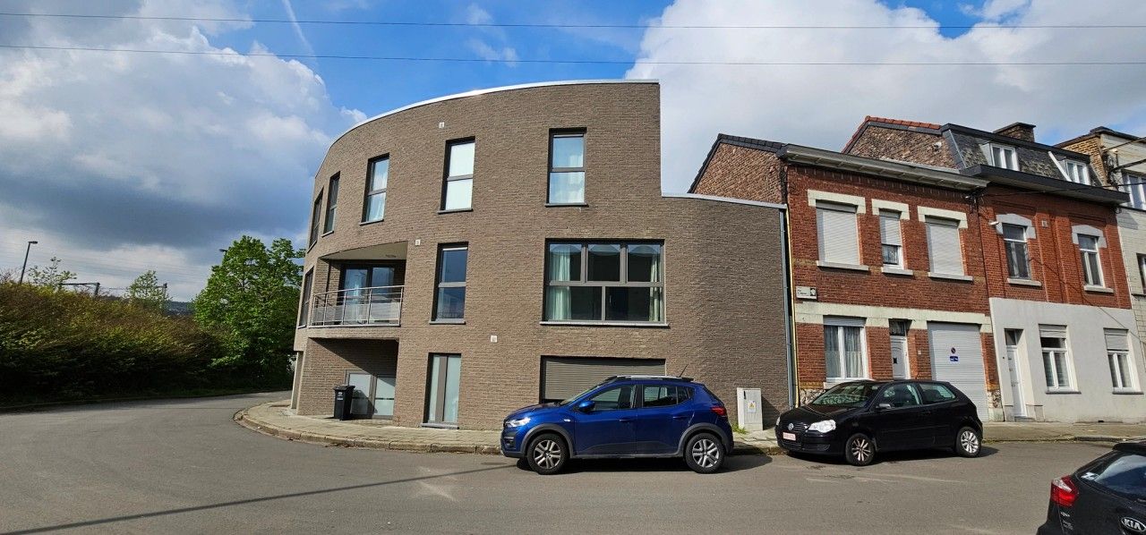 Vue de l’entrée de l’immeuble résidentiel à Flémalle, avec façade courbe en briques, balcon couvert et larges ouvertures des appartements.