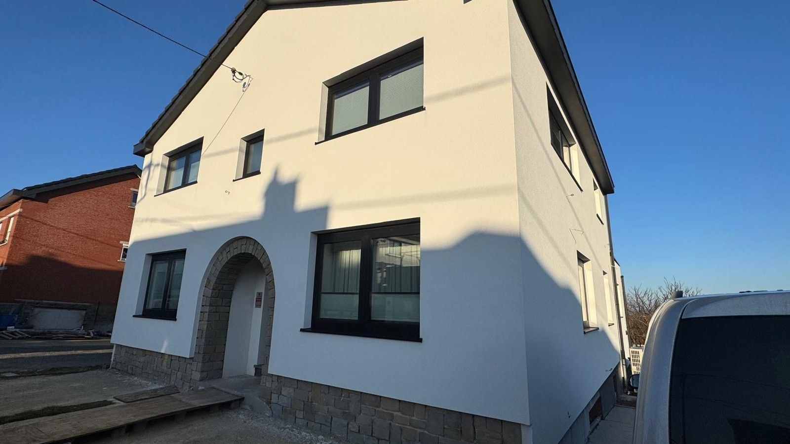 Vue de la façade avant rénovée de l’habitation à Vottem, avec enduit blanc, châssis foncés, soubassement en pierre et entrée cintrée conservée.